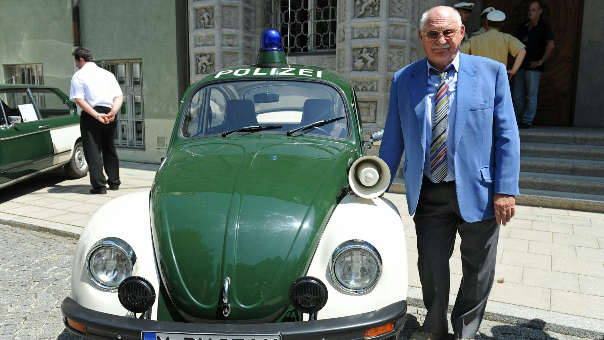 Schauspieler Rolf Schimpf steht vor dem Polizeipräsidium neben einem alten VW-Käfer-Polizeiauto.