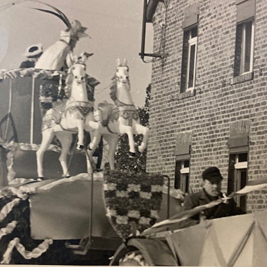 Eine Schwarz-Weiß-Aufnahme zeigt einen Karnevalswagen der KG Vernich mit Pferdefiguren, der an einem Backsteinhaus vorbeifährt.