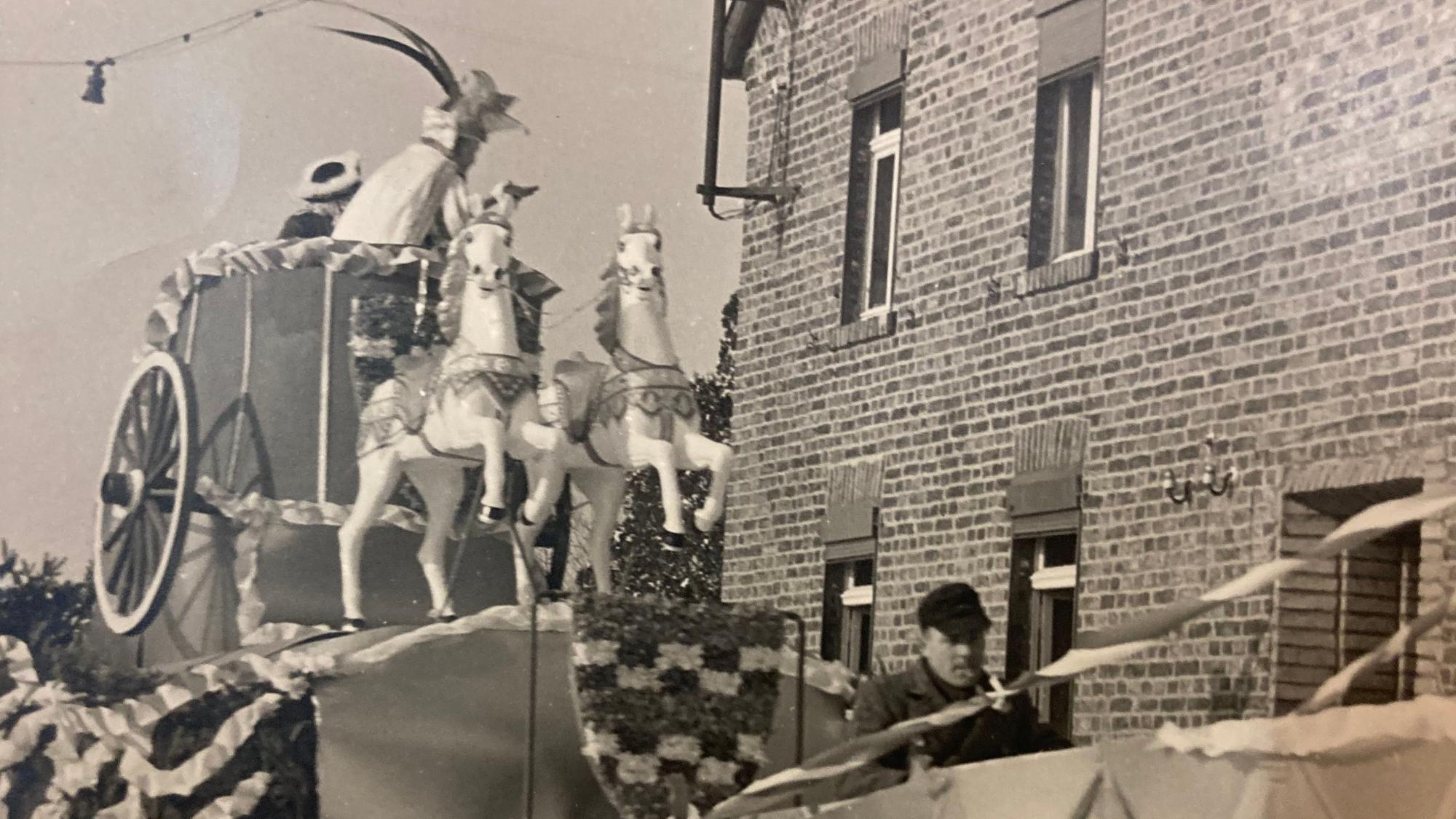 Eine Schwarz-Weiß-Aufnahme zeigt einen Karnevalswagen der KG Vernich mit Pferdefiguren, der an einem Backsteinhaus vorbeifährt.