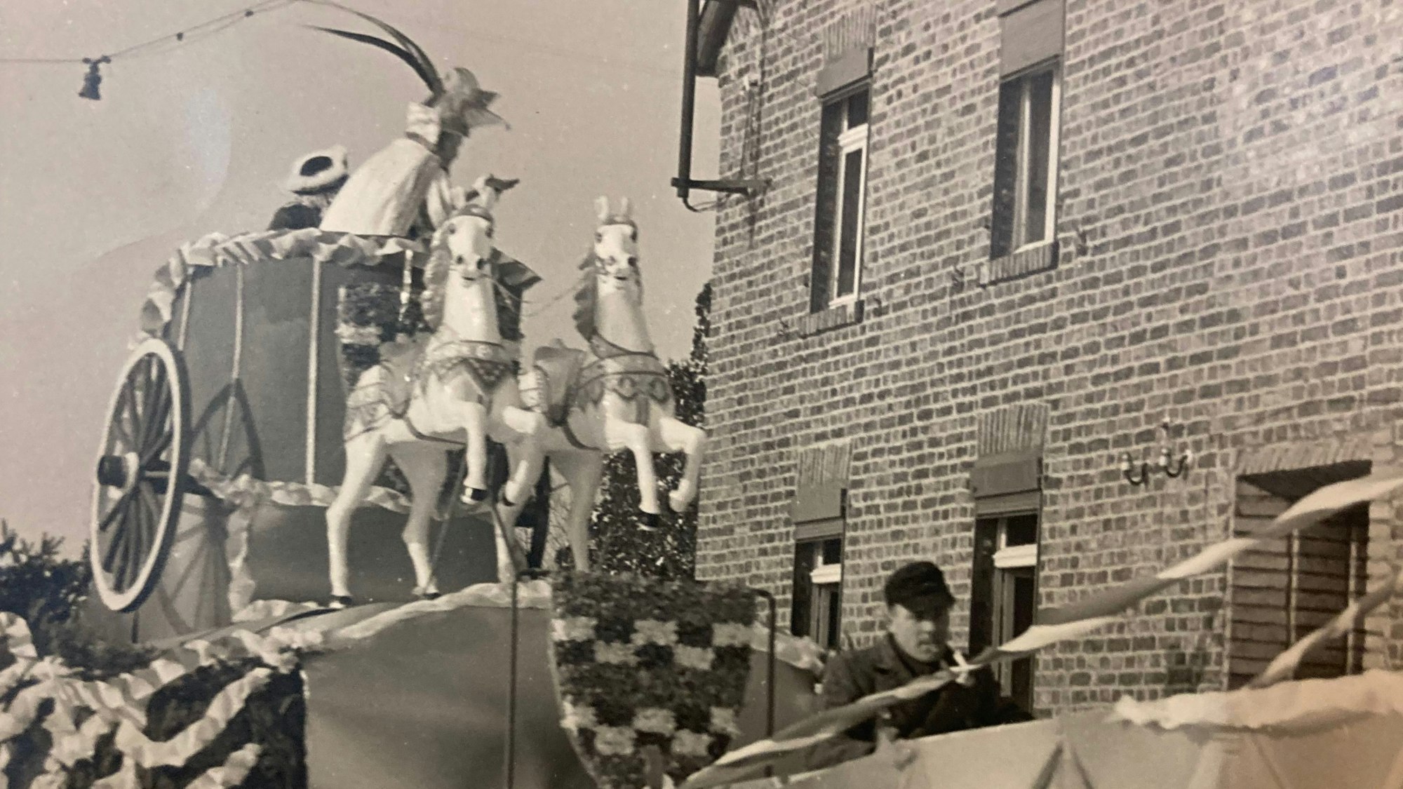 Eine Schwarz-Weiß-Aufnahme zeigt einen Karnevalswagen der KG Vernich mit Pferdefiguren, der an einem Backsteinhaus vorbeifährt.