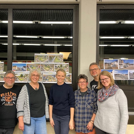 Sieben Menschen stehen fröhlich vor Glasscheiben, an denen Bilder hängen.