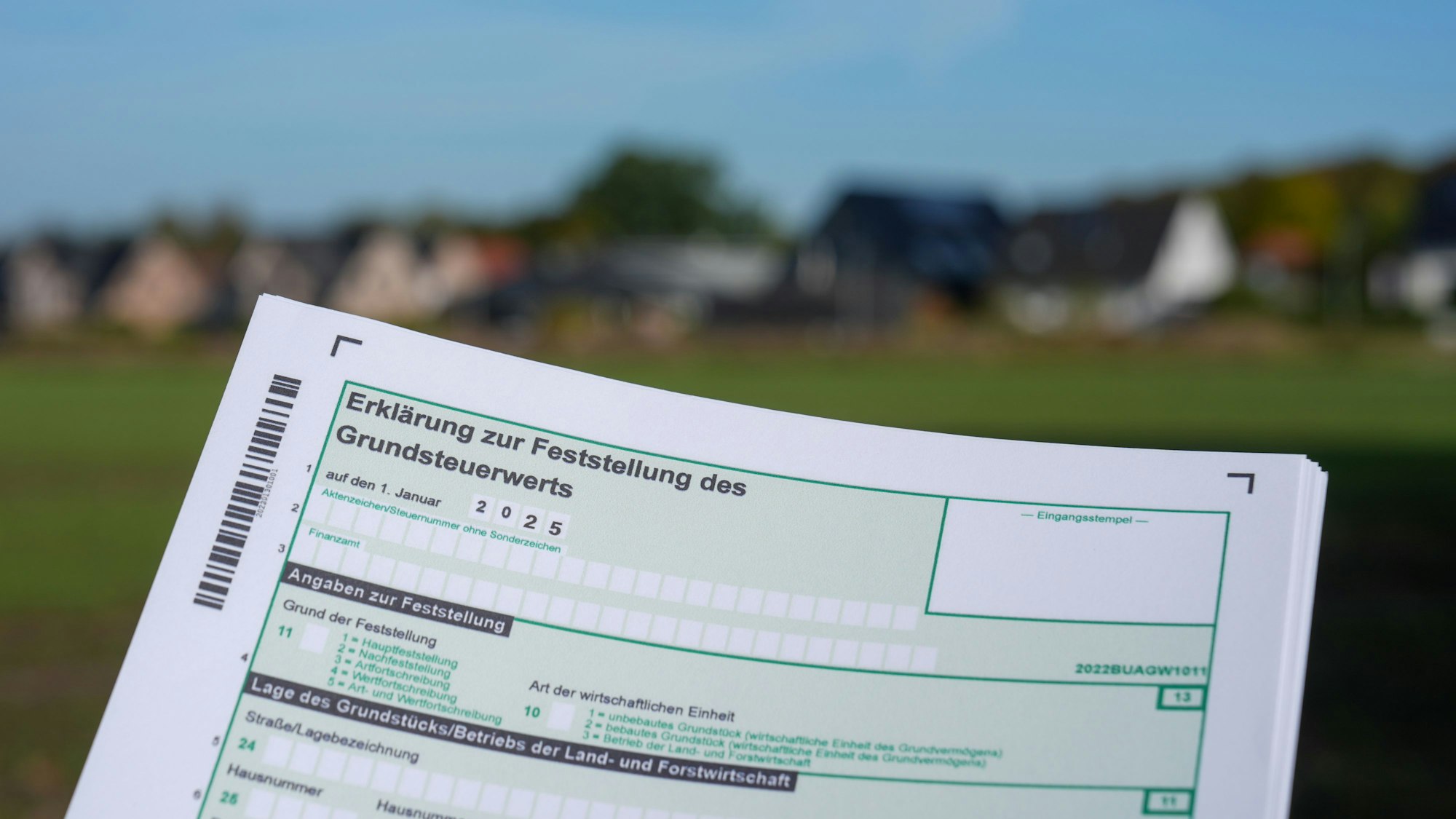 Ein Formular mit der Erklärung zur Feststellung des Grundsteuerwerts für das Jahr 2025 wird vor eine Wohnsiedlung hochgehalten.