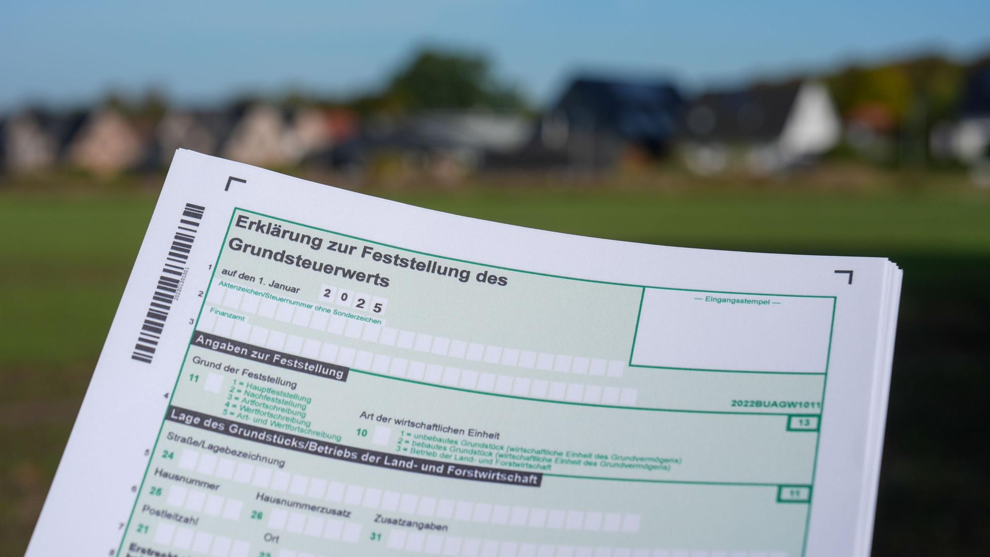 Eine Erklärung zur Feststellung des Grundsteuerwerts für das Jahr 2025 wird vor eine Wohnsiedlung hochgehalten.