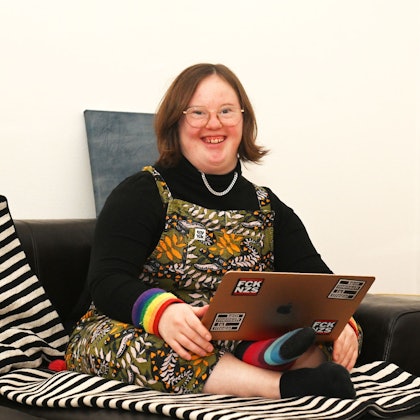 Eine junge Frau sitzt im Schneidersitz auf dem Sofa, auf den Knien hat sie einen Laptop.