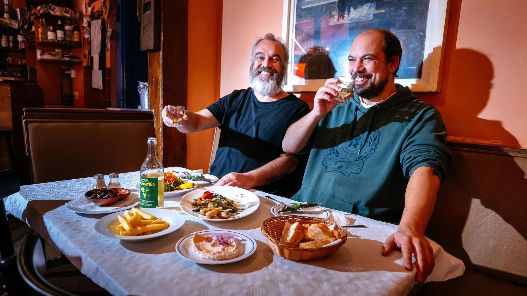 Zwei Männer sitzen an einem gedeckten Tisch und haben ein kleines Gläschen in der Hand.
