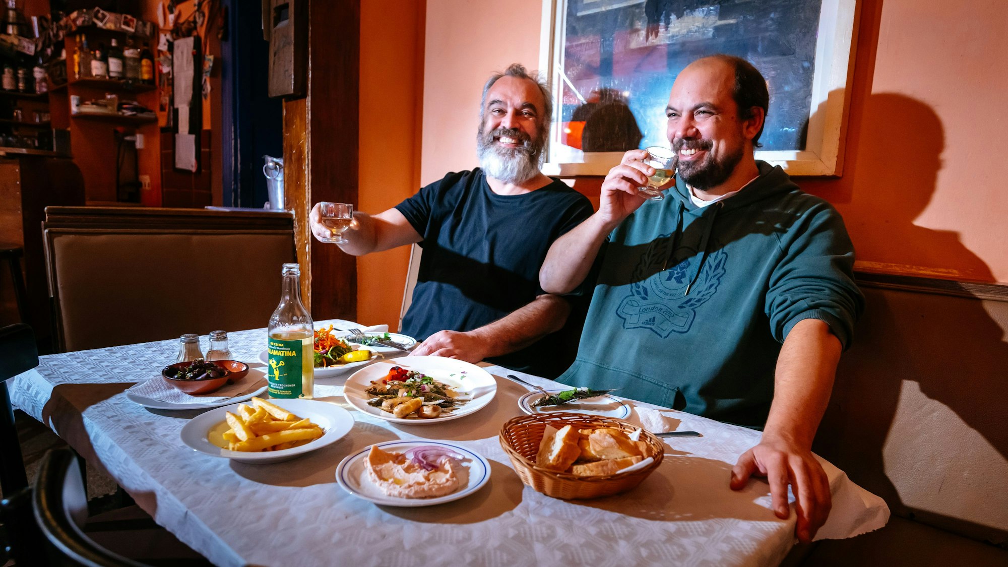 Zwei Männer sitzen an einem gedeckten Tisch und haben ein kleines Gläschen in der Hand.