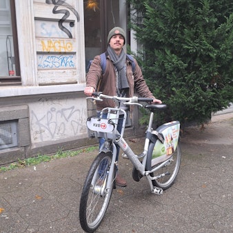 Fabio Botero steht mit einem KVB-Rad auf dem Gehweg.