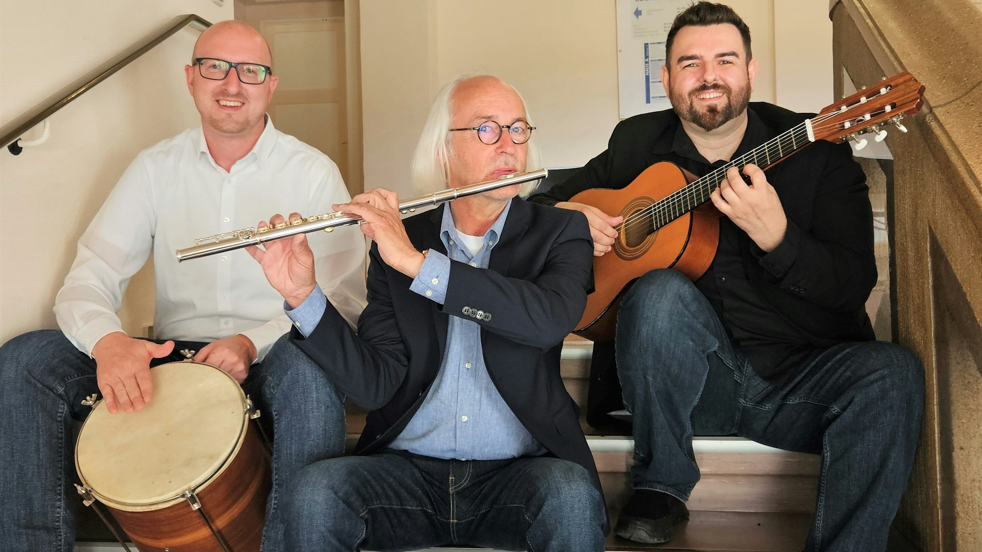 Drei Männer sitzen auf einer Treppe, einer spielt Trommel, einer Querflöte und einer Gitarre.