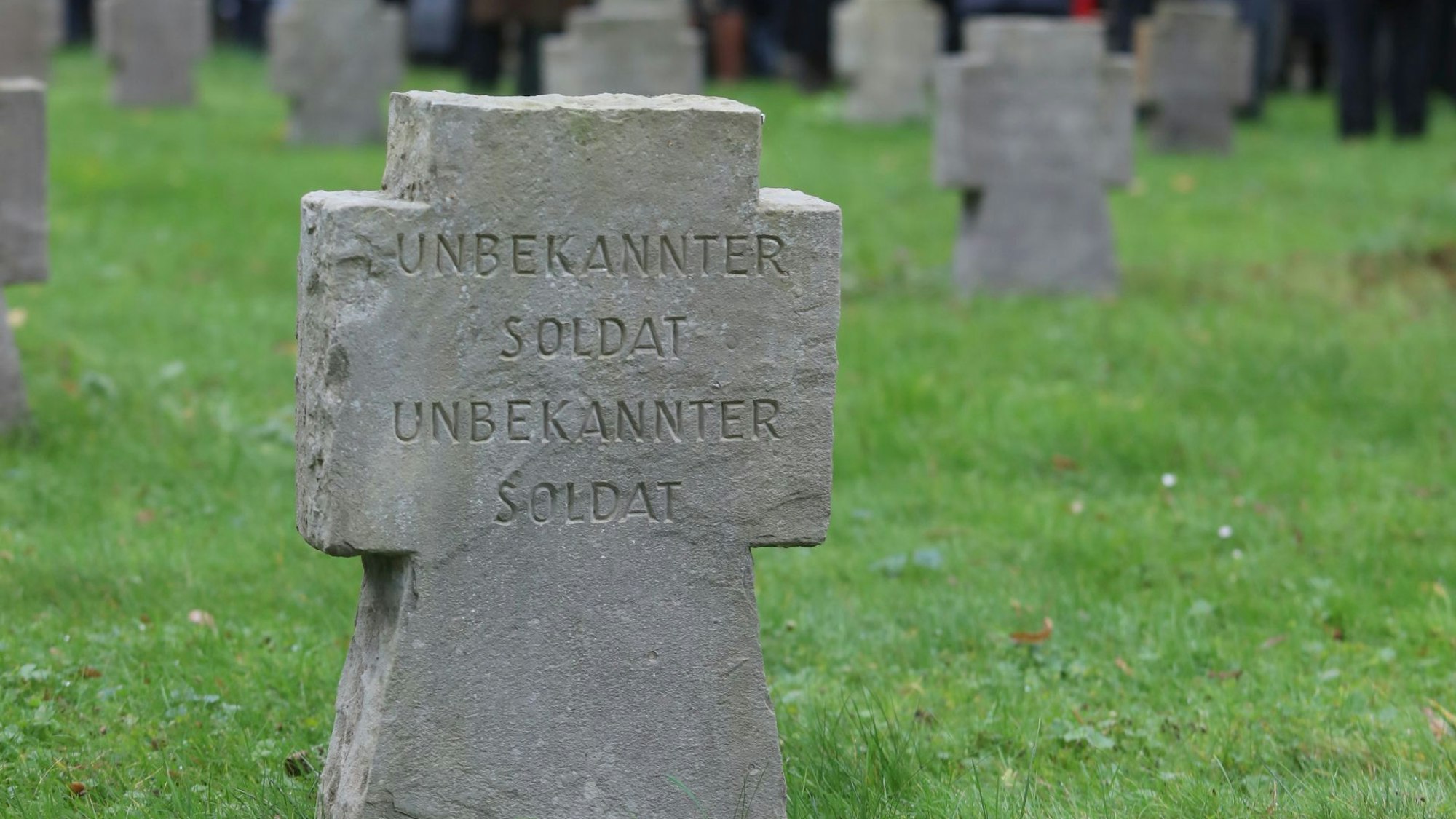 Eine Wiese mit Kriegsgräbern. Auf einem Stein in Kreuzform steht „Unbekannter Soldat“.