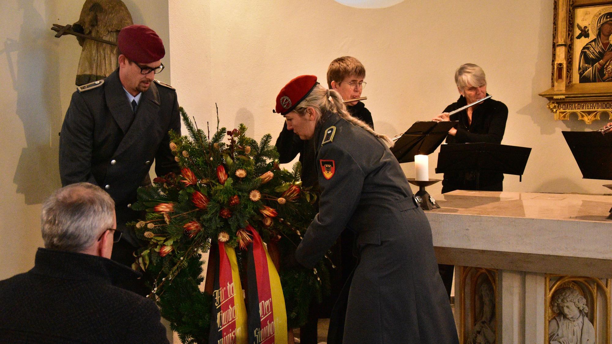 Ein Soldat und eine Soldatin legen einen Kranz mit Blumen und Schleifen in den Nationalfarbe Schwarz, Rot, Gold nieder. Im Hintergrund spielen zwei Personen Querflöte.