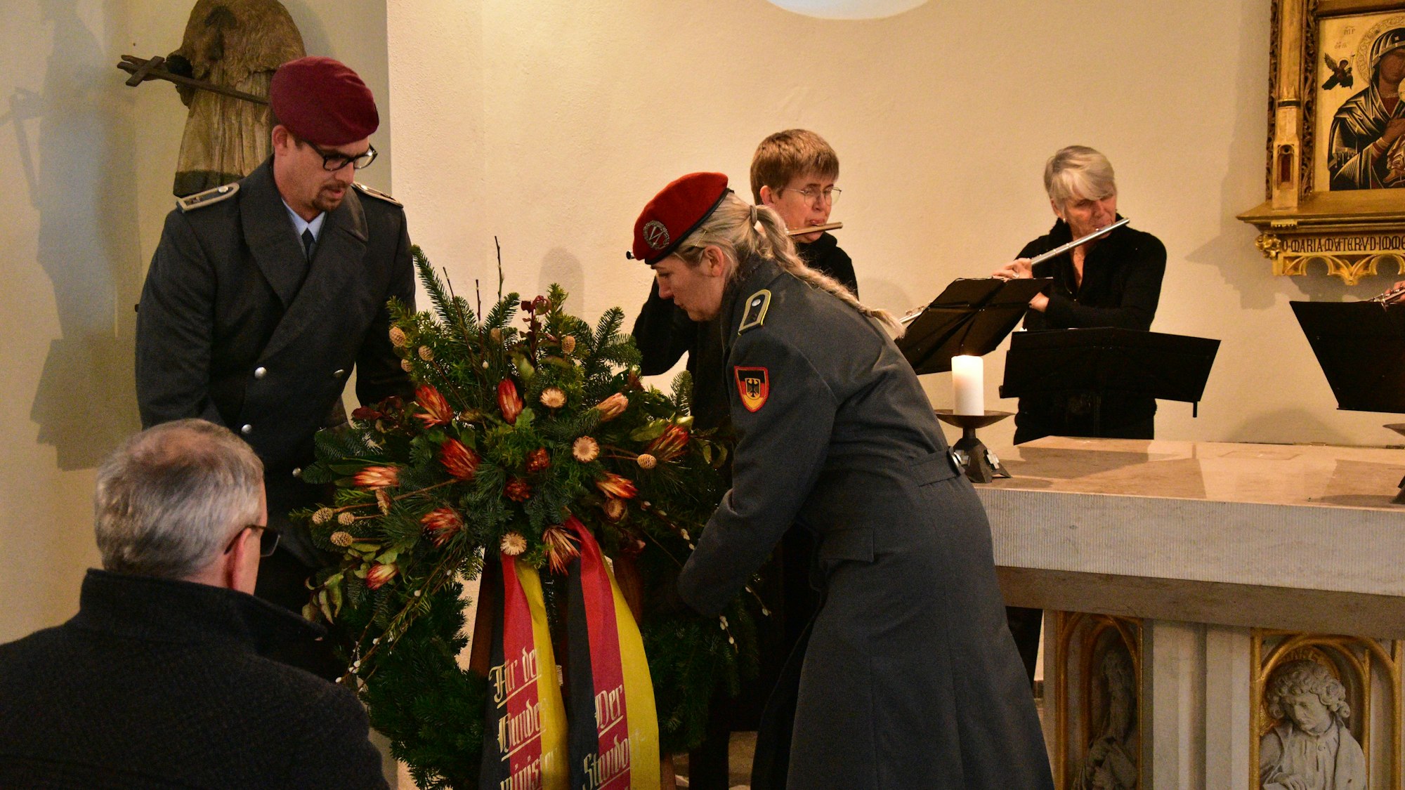 Ein Soldat und eine Soldatin legen einen Kranz mit Blumen und Schleifen in den Nationalfarbe Schwarz, Rot, Gold nieder. Im Hintergrund spielen zwei Personen Querflöte.