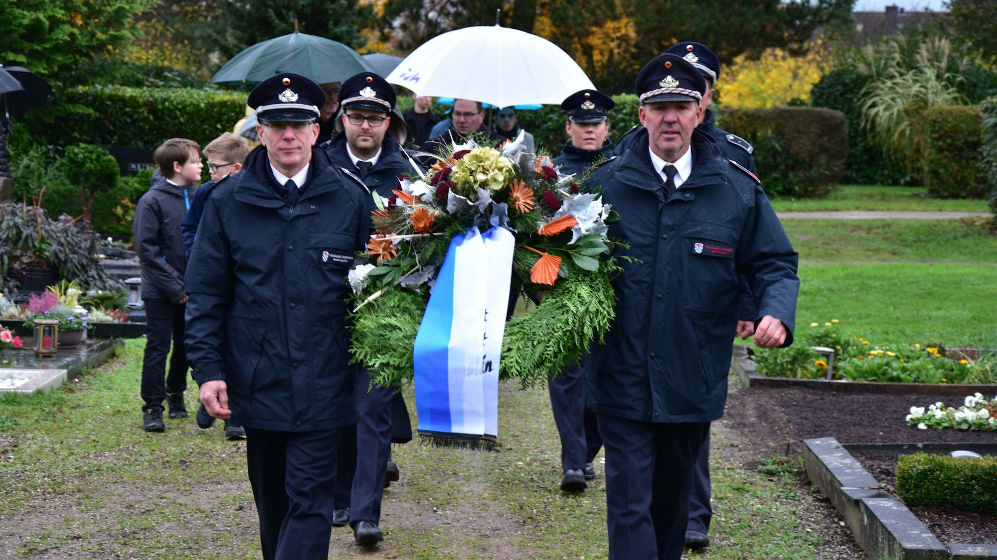 Zwei Männer mit Schirmmützen tragen auf einem Friedhof einen Kranz mit Blumenschmuck und blau-weißen Schleifen. Es folgen Menschen mit Regenschirmen.