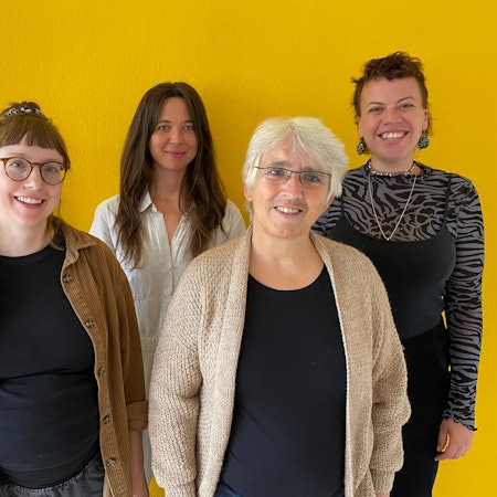 Fünf Frauen unterschiedlichen Alters stehen vor einer gelben Wand und lächeln in die Kamera.