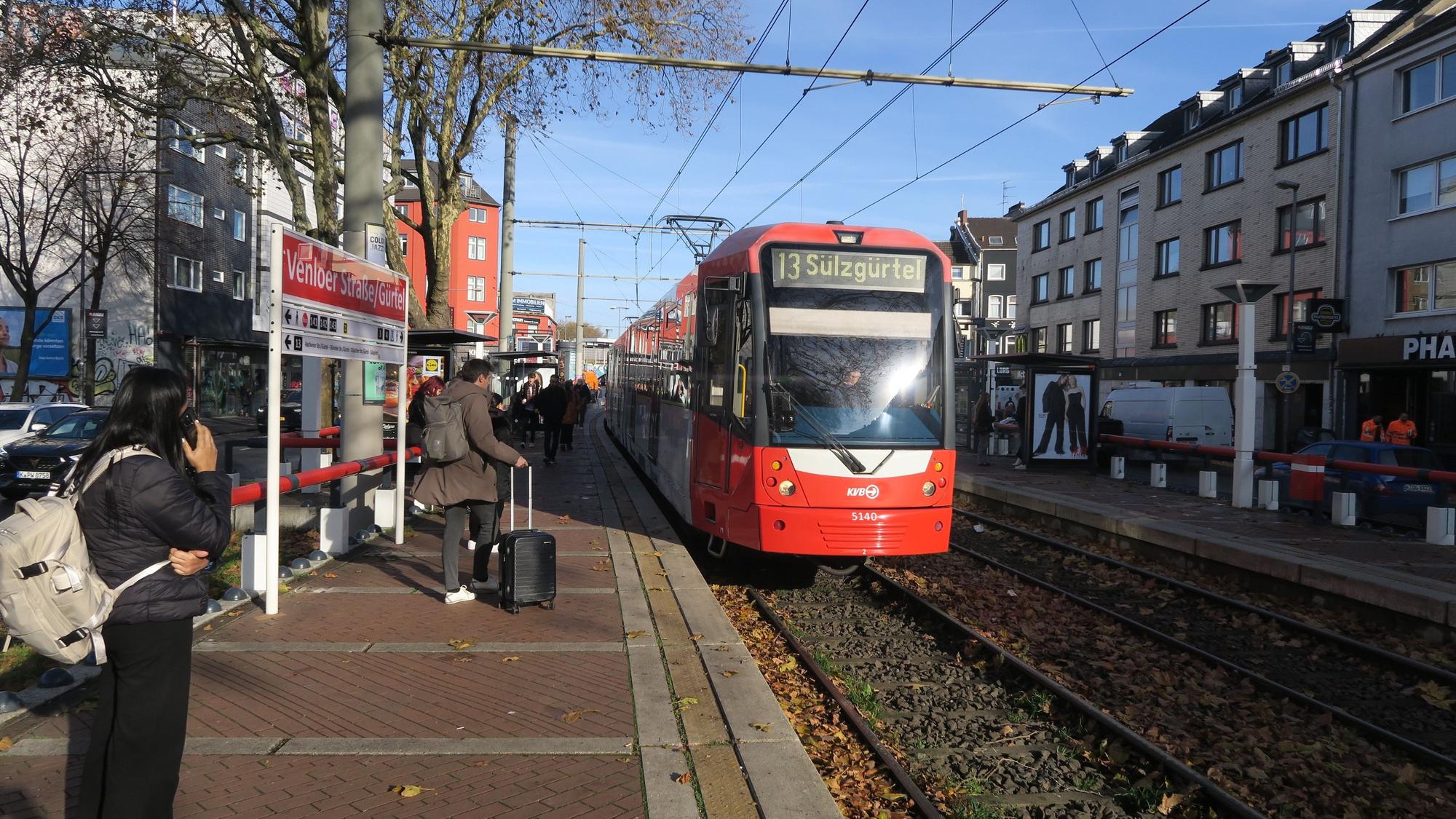 An einem Bahnsteig stehen eine Frau und ein Mann, ein Zug der Linie 13 fährt gerade ein.