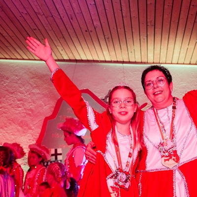 Eine Frau und ein Mädchen mit Karnevalsorden haben die Arme umeinander gelegt und strecken die freien Arme in die Luft.