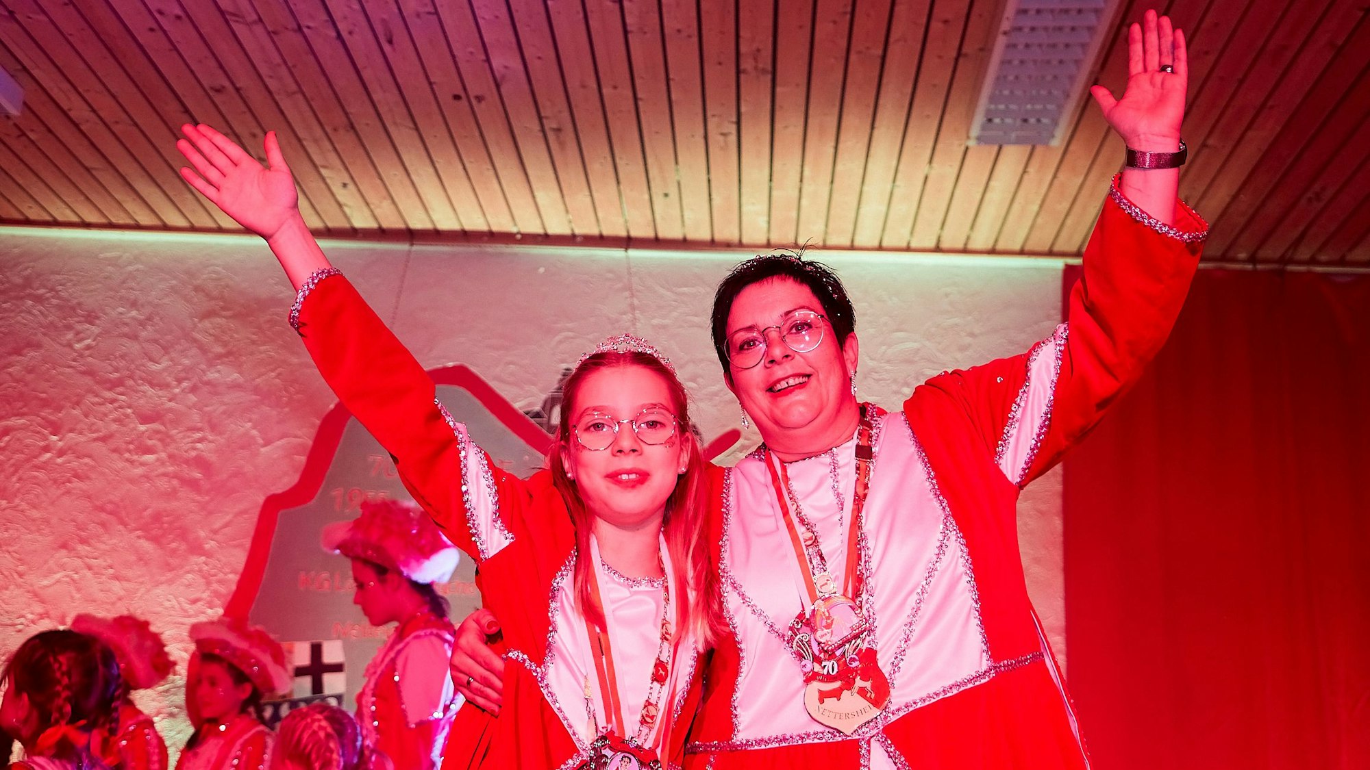 Eine Frau und ein Mädchen mit Karnevalsorden haben die Arme umeinander gelegt und strecken die freien Arme in die Luft.