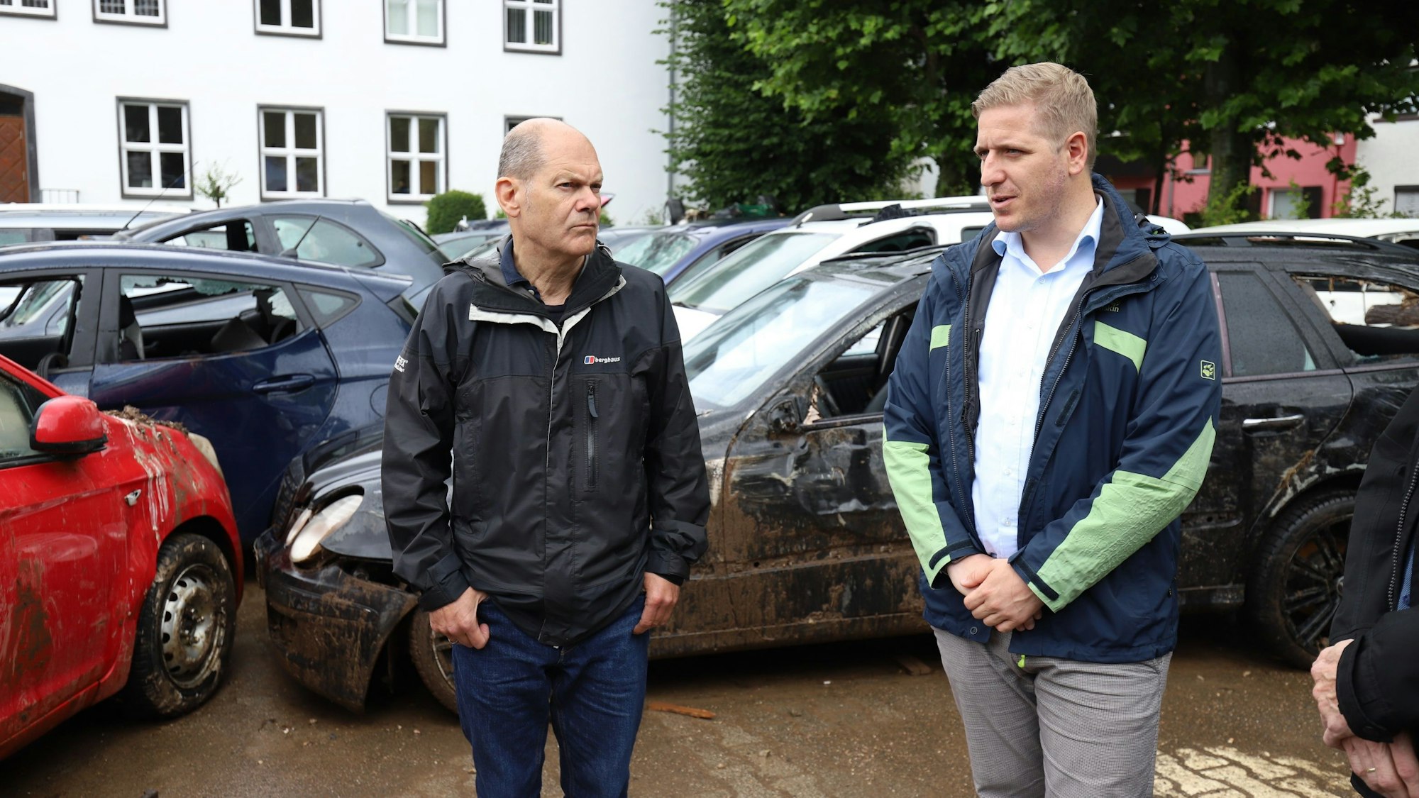Olaf Scholz und Markus Ramers tragen beide Regenjacken. Im Hintergrund sind bei der Flut zerstörte Autos zu sehen.