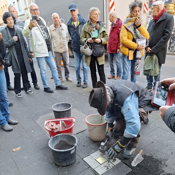 Ein Mann mit Hut arbeitet an einem Bürgersteig. Er ist dabei, einen glänzenden Erinnerungsstein zu verlegen.