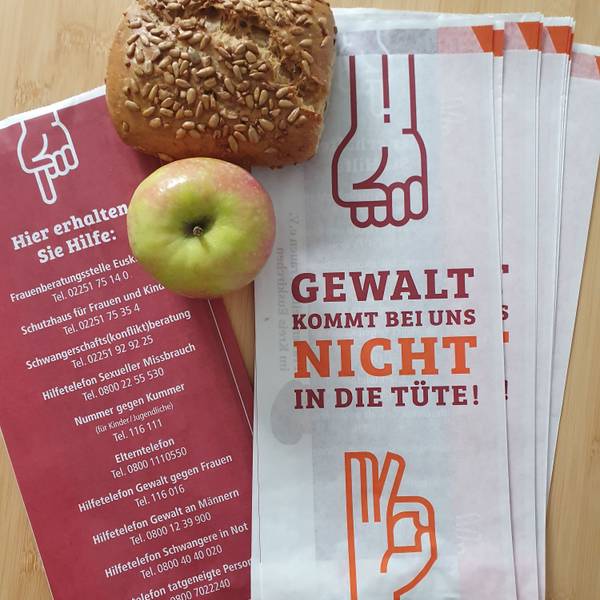 Mehrere Brötchentüten liegen auf einem Tisch, daneben ein Apfel und ein Körnerbrötchen. Auf der Vorderseite der Tüten steht „Gewalt kommt bei uns nicht in die Tüte“, auf der Rückseite sind Telefonnummern verschiedener Anlaufstellen, falls man von Gewalt betroffen ist.