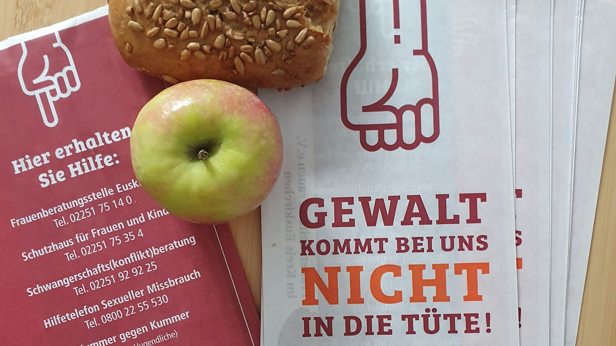 Mehrere Brötchentüten liegen auf einem Tisch, daneben ein Apfel und ein Körnerbrötchen. Auf der Vorderseite der Tüten steht „Gewalt kommt bei uns nicht in die Tüte“, auf der Rückseite sind Telefonnummern verschiedener Anlaufstellen, falls man von Gewalt betroffen ist.