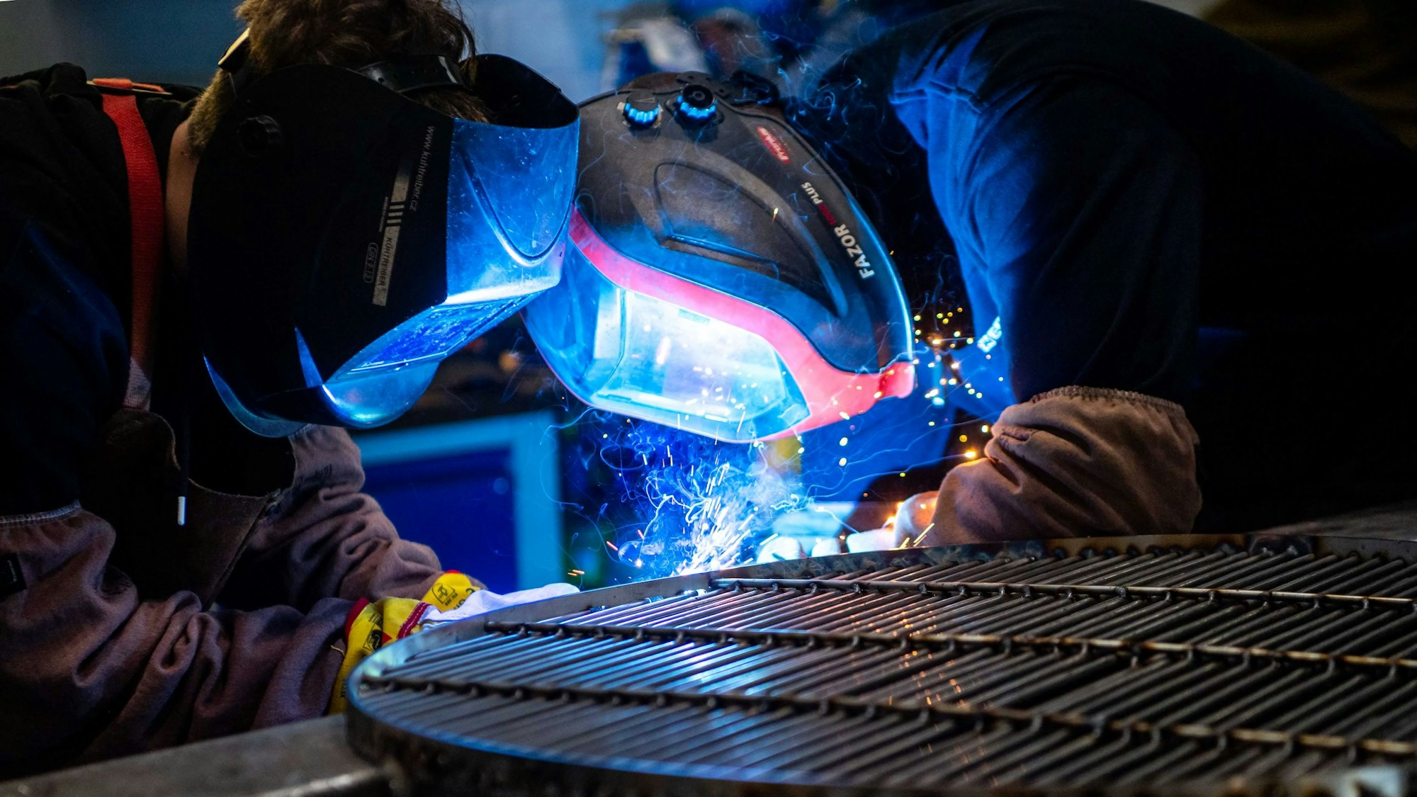 Zwei Jugendliche stehen mit Schutzmasken an einem Eisengitter und schweißen etwas. Ihre Gesichter sind in blaues Licht getaucht und es sprühen Funken.