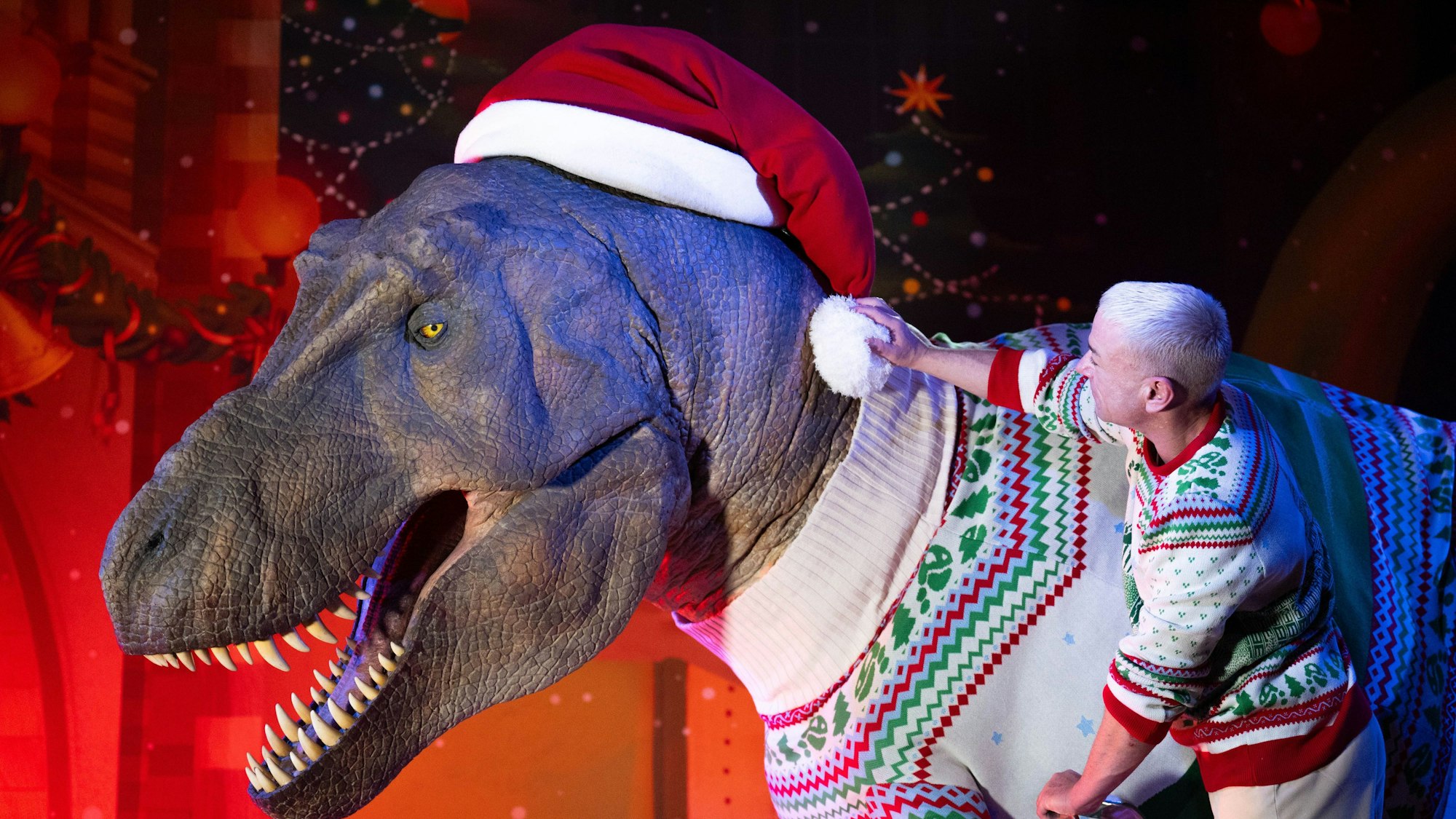 Ein Mitarbeiter rückt eine riesige Weihnachtsmannmütze auf einem Raubsaurier zurecht, der im Natural History Museum bereits einen Weihnachtspulli trägt.