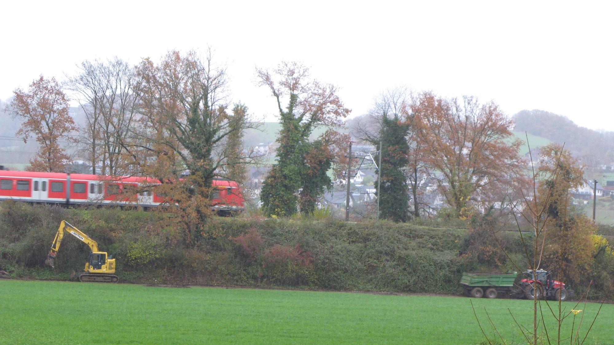 Ein Bagger steht vor einem Bahndamm, auf dem ein S-Bahnzug fährt. Ein Traktor zieht einen beladenen Anhänger weg.