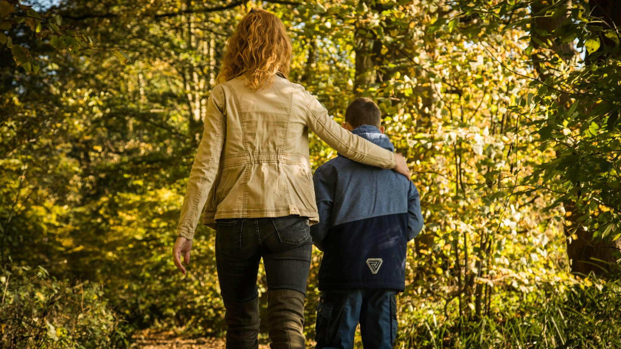 Eine Frau hält einen Arm um einen Jungen und geht mit ihm durch den Wald.
