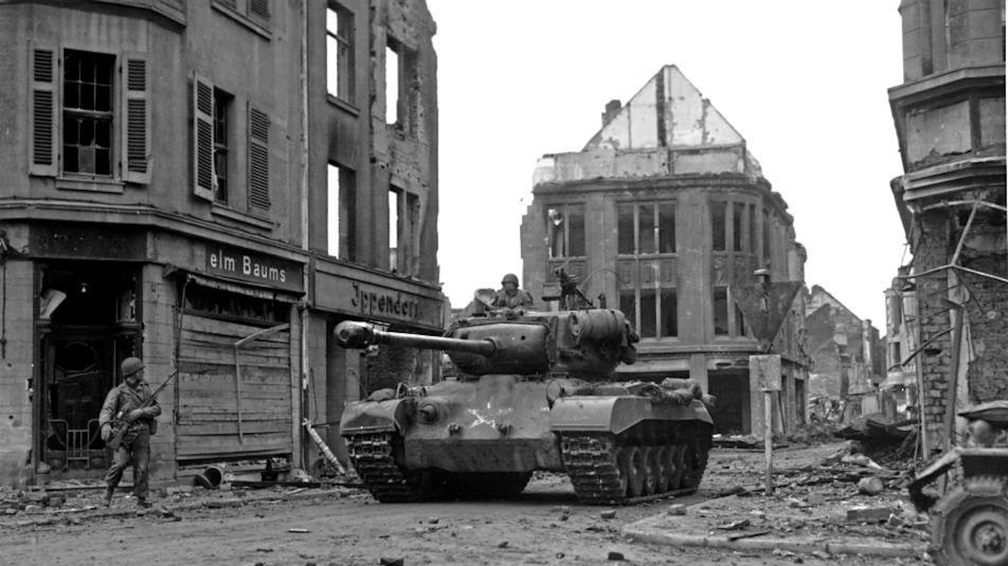 Eine historische Aufnahme zeigt einen Panzer, umgeben von zerstörten Häusern. Am Straßenrand steht ein Soldat.