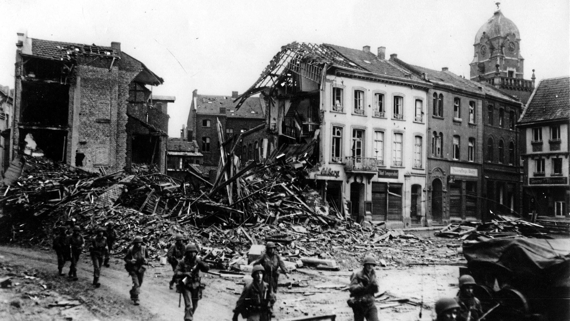 Das Schwarz-Weiß-Bild zeigt Soldaten in Zweierreihen, im Hintergrund zerstörte Gebäude.