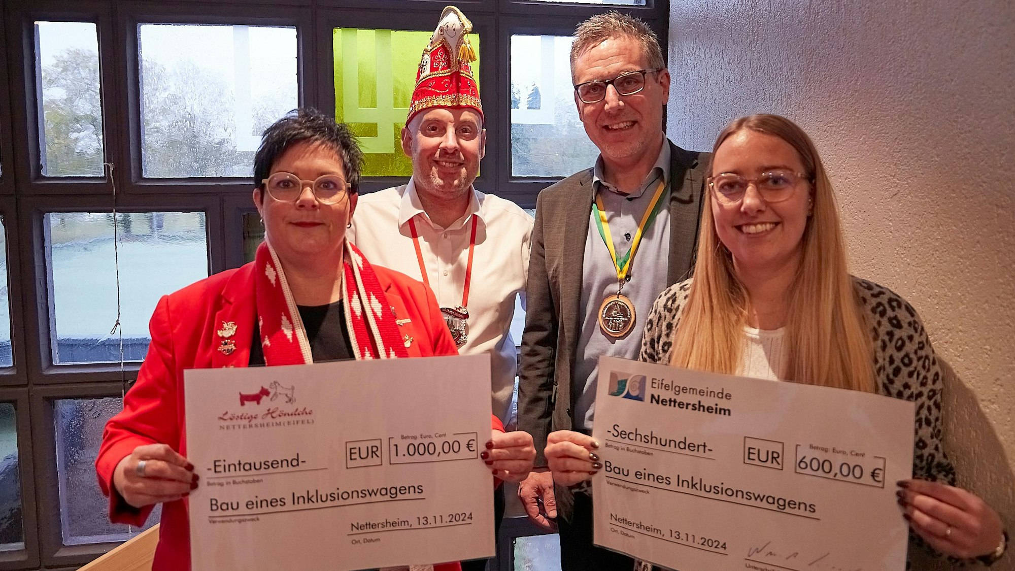 Zwei Frauen und zwei Männer stehen vor einem Fenster und halten zwei Schecks in die Kamera.