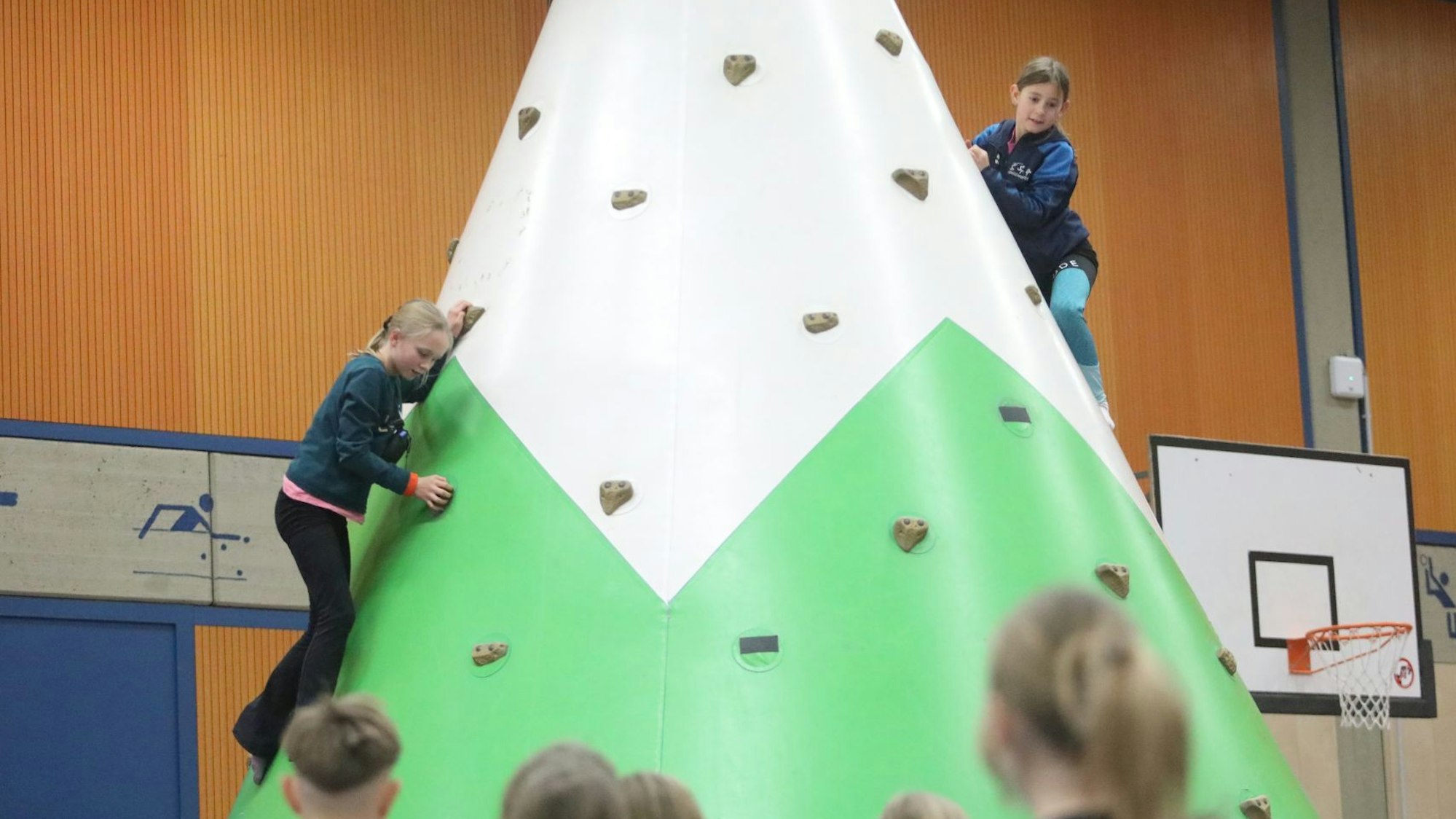 Zwei Mädchen klettern an einem Kletterfelsen, mehrere Kinder schauen zu.