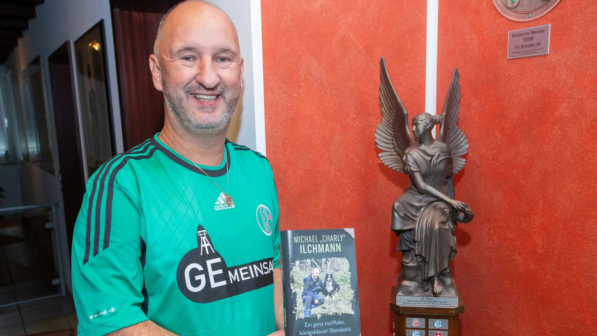 Foto eines Fußballfans vor einer Statue, in der Hand hält er ein Buch.