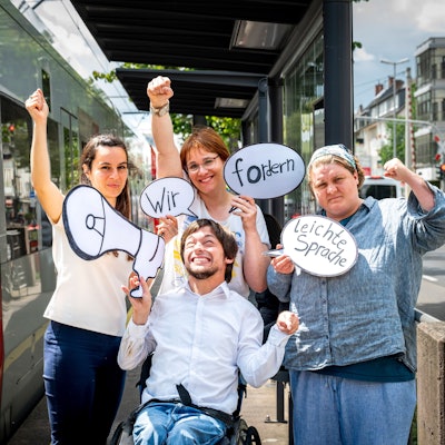 Auf dem Bild stehen Margarita Lüdtke Miró (v. l). Maximilian Westphal (vorne), Silke Degwitz und Dinah Köppen an einer KVB-Haltestelle. Sie halten Protestplakate.