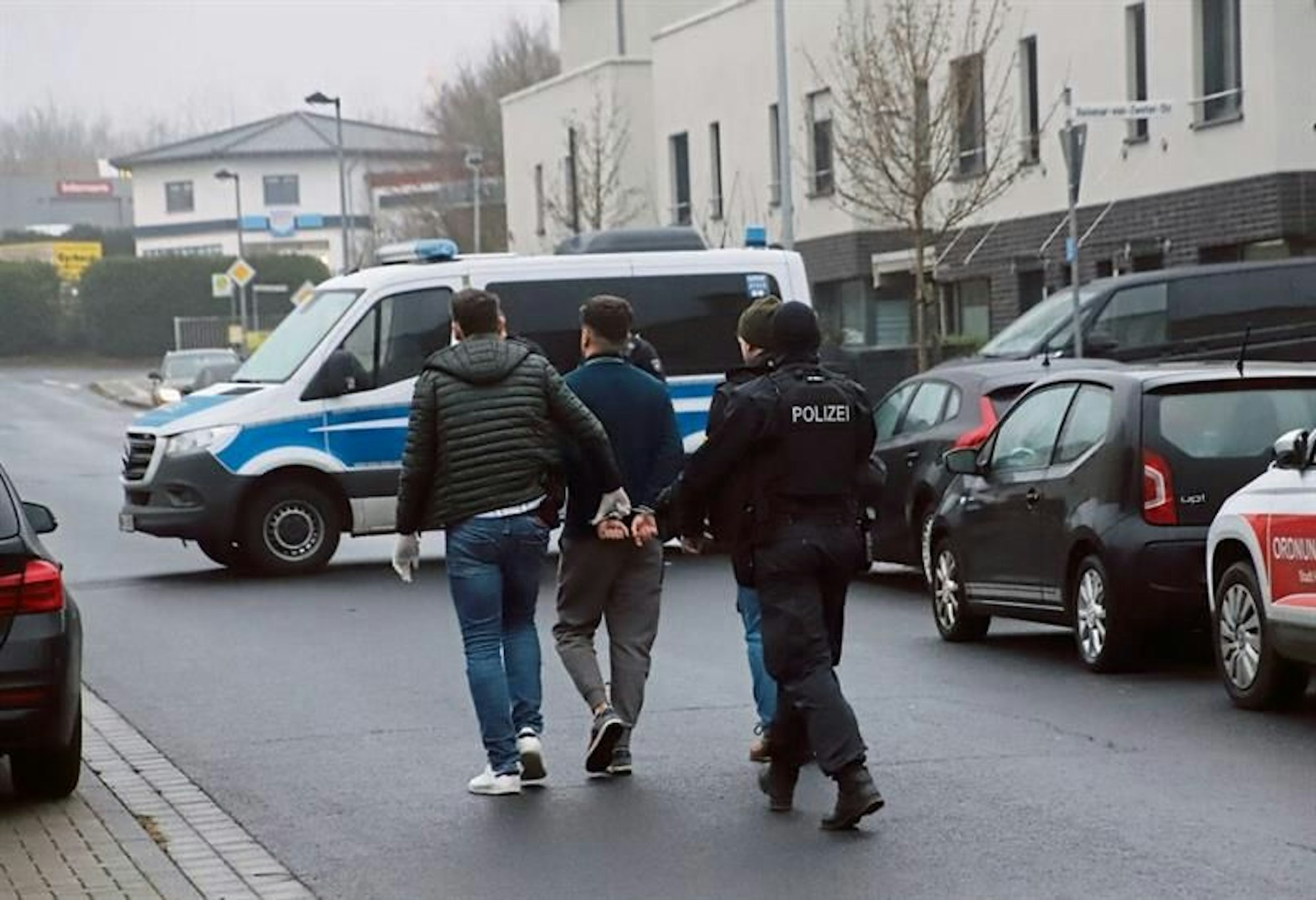 Ein Mann mit auf dem Rücken gefesselten Händen wird von mehreren Männern abgeführt, einer trägt eine Weste mit dem Aufdruck "Polizei". Ein Polizeitransporter steht quer auf der Straße.
