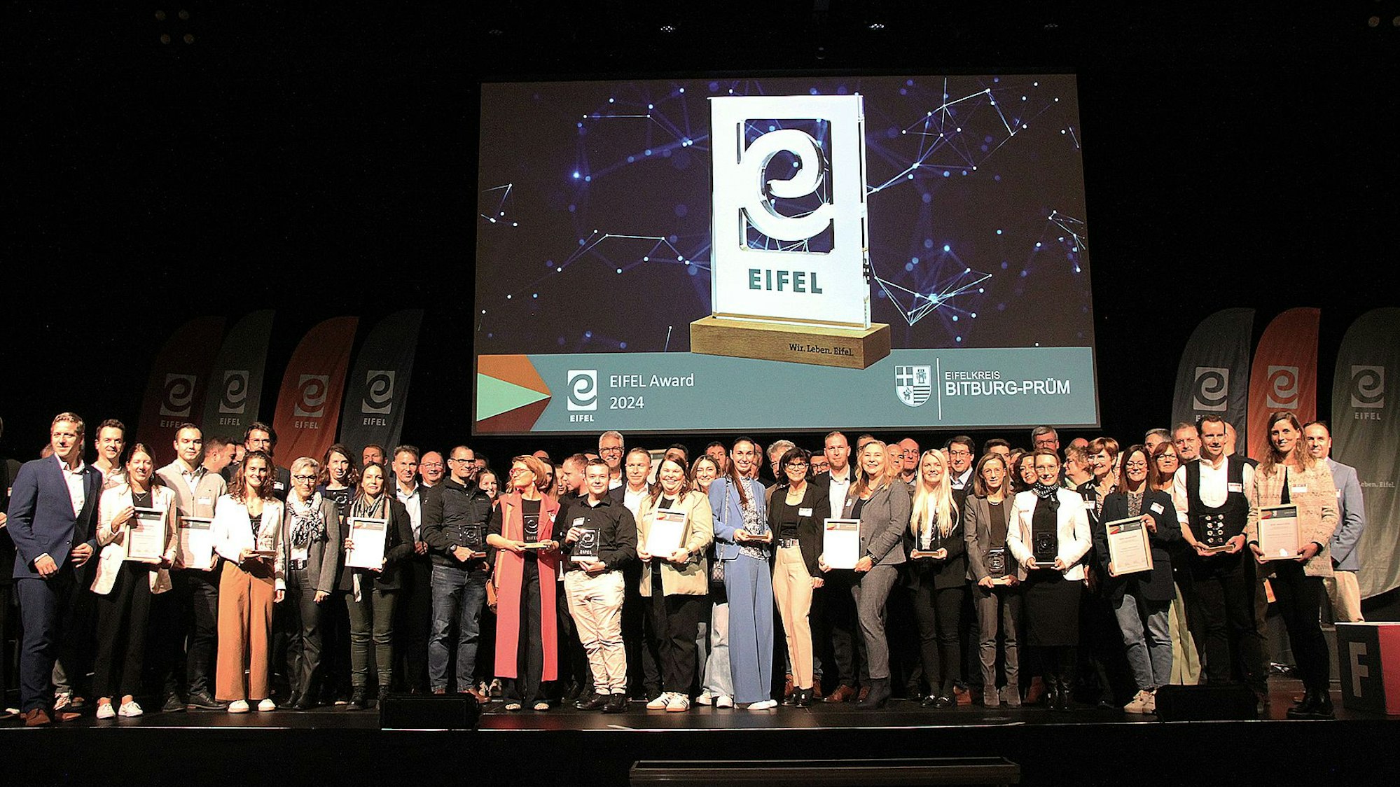 Auf einer Bühne stehen viele Menschen dicht bei andern. Im Hintergrund zeigt ein Bildschirm den Preis der Eifel Awards.
