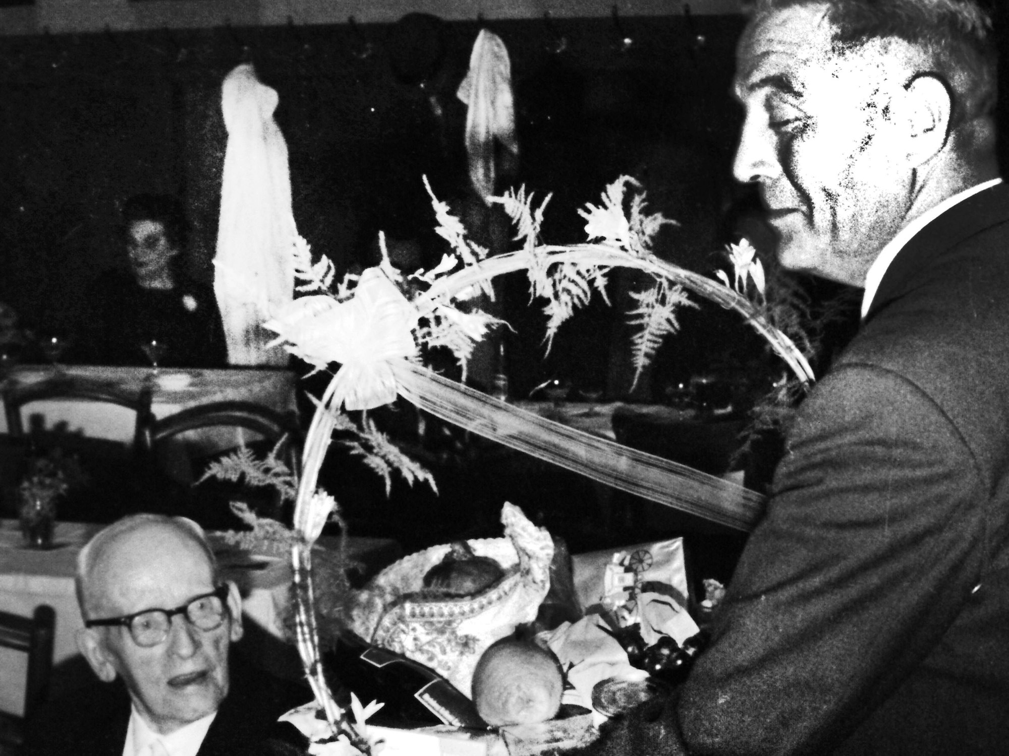 Ein Mann im Anzug überreicht einem älteren Herrn mit Brille, der sitzt, einen Präsentkorb.