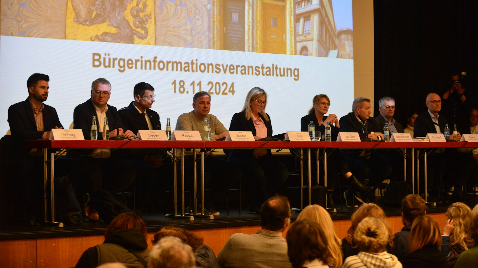 Auf einem Podium sitzen mehrere Frauen und Männer hinter Tischen, darauf stehen ihre Namensschilder.