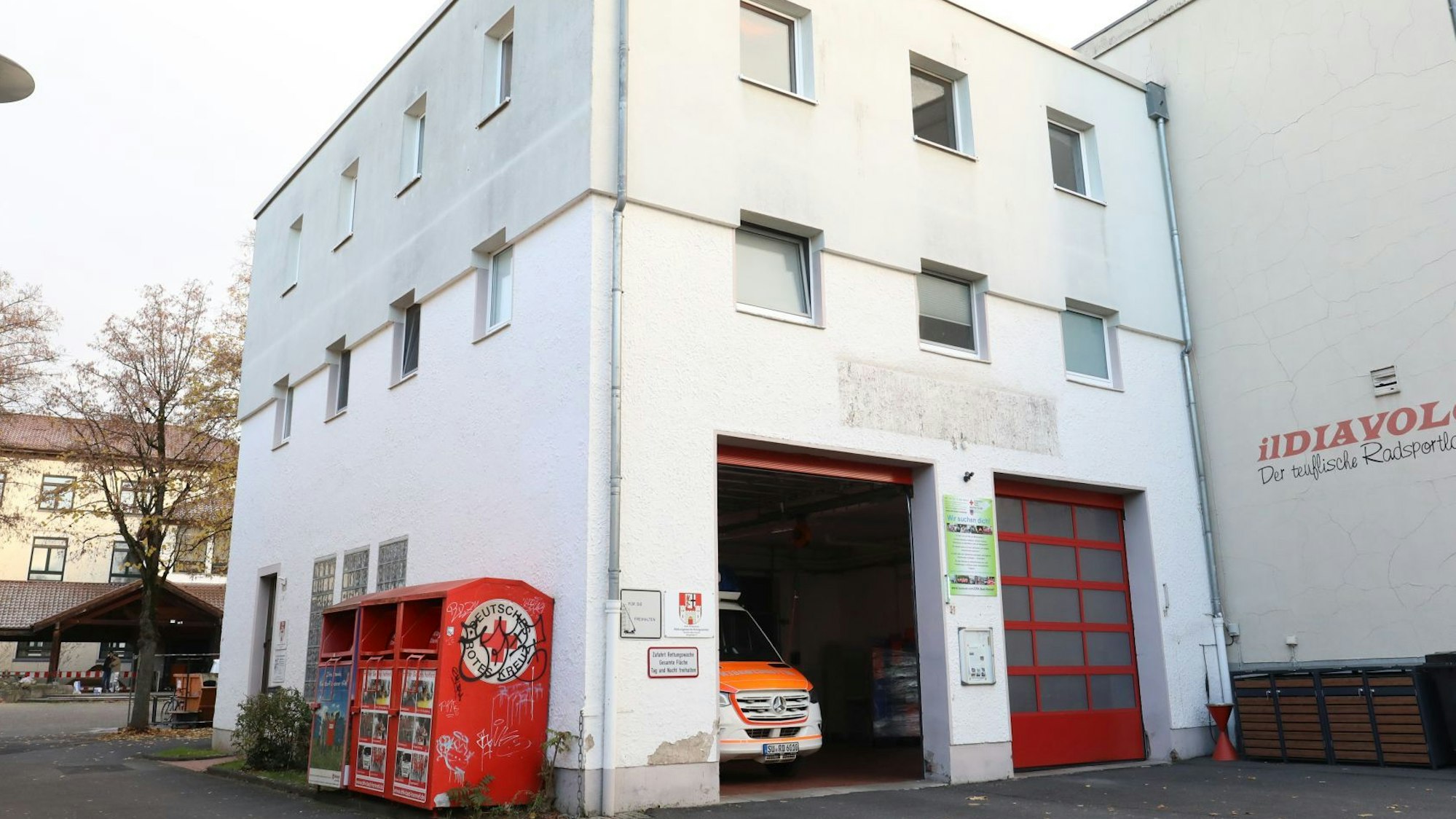 Ein weißes Gebäude mit zwei roten Rolltoren; eines steht offen, ein Rettungswagen ist zu sehen.