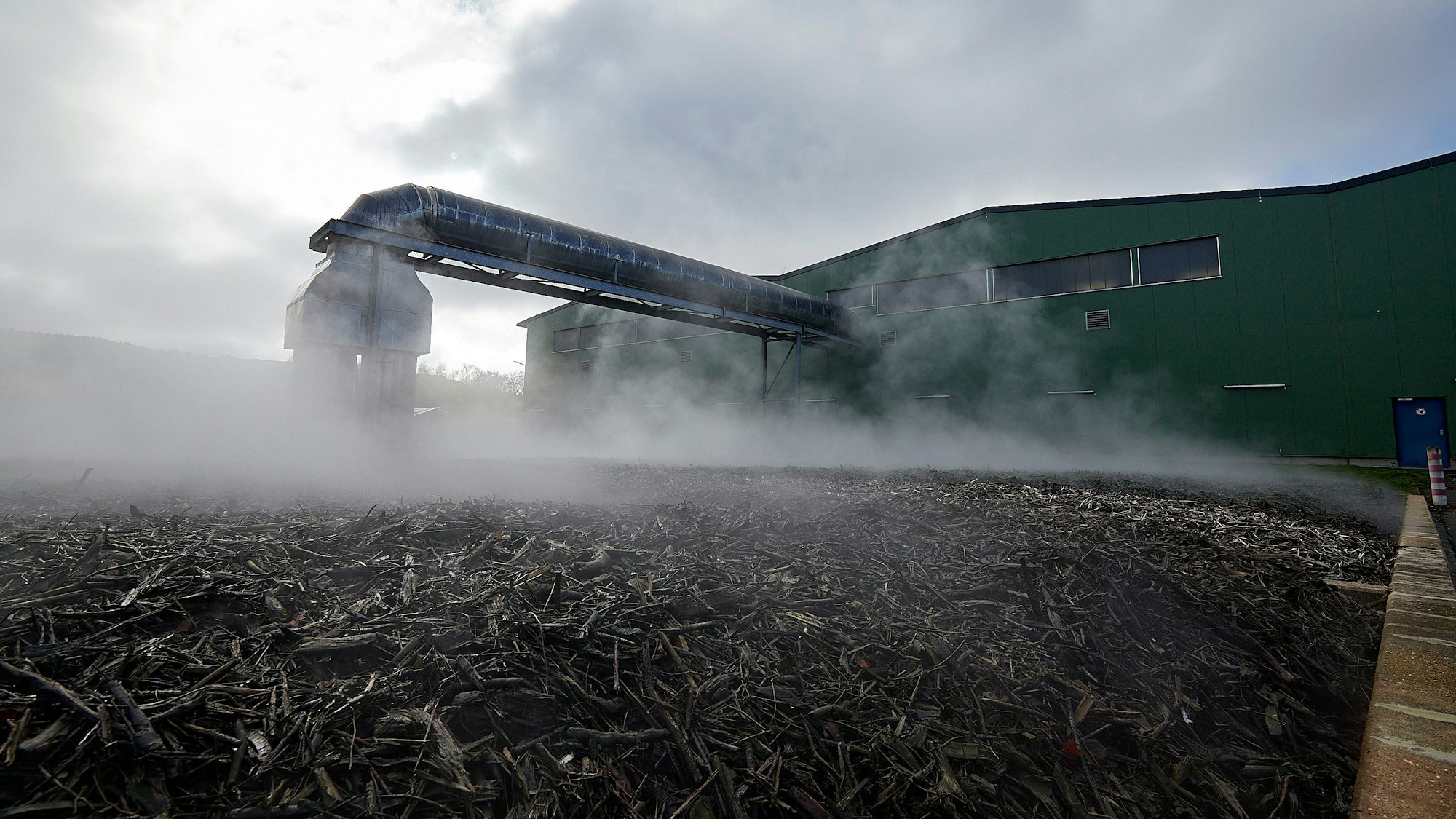 Bei einer Kompostieranlage liegt Strauch und Astwerk auf dem Boden. Dampf steigt im Hintergrund auf.