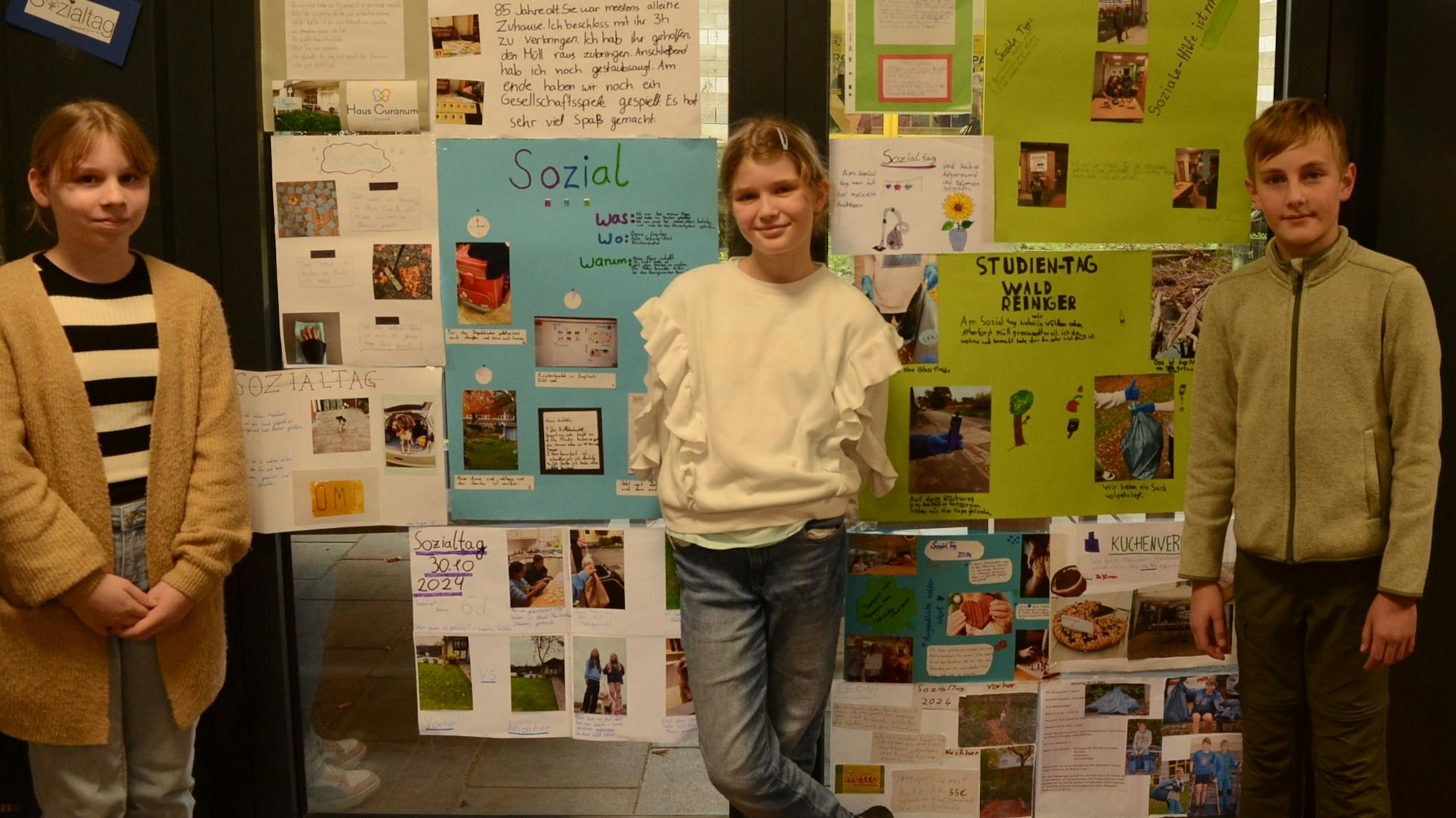 Zwei Mädchen und ein Junge stehen vor einer Fensterwand mit Collagen.