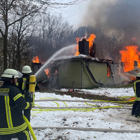 Feuerwehrleute löschen eines der langgestreckten Gebäude in der ZUE in Vogelsang. Die Flammen lodern aus dem Dach.