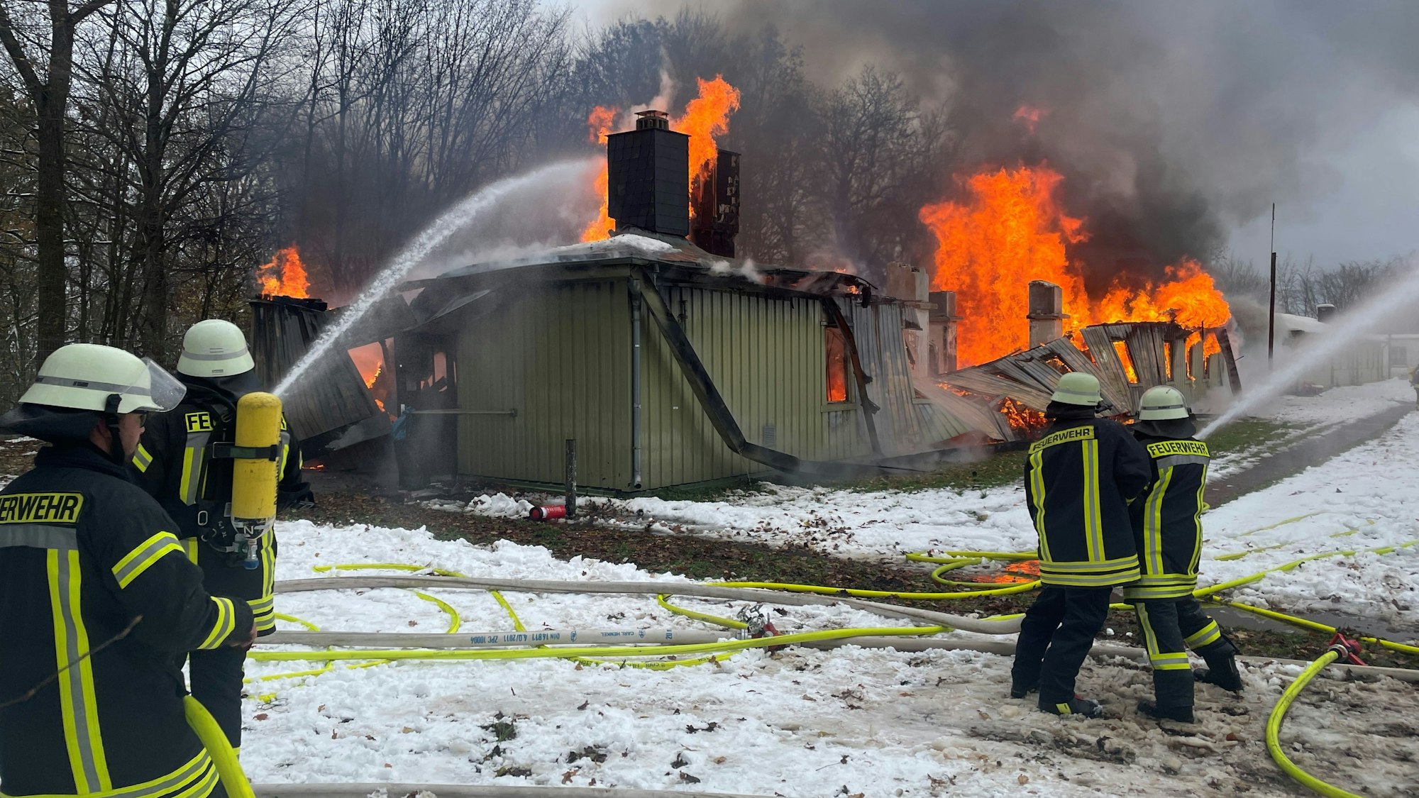 Feuerwehrleute löschen eines der langgestreckten Gebäude in der ZUE in Vogelsang. Die Flammen lodern aus dem Dach.