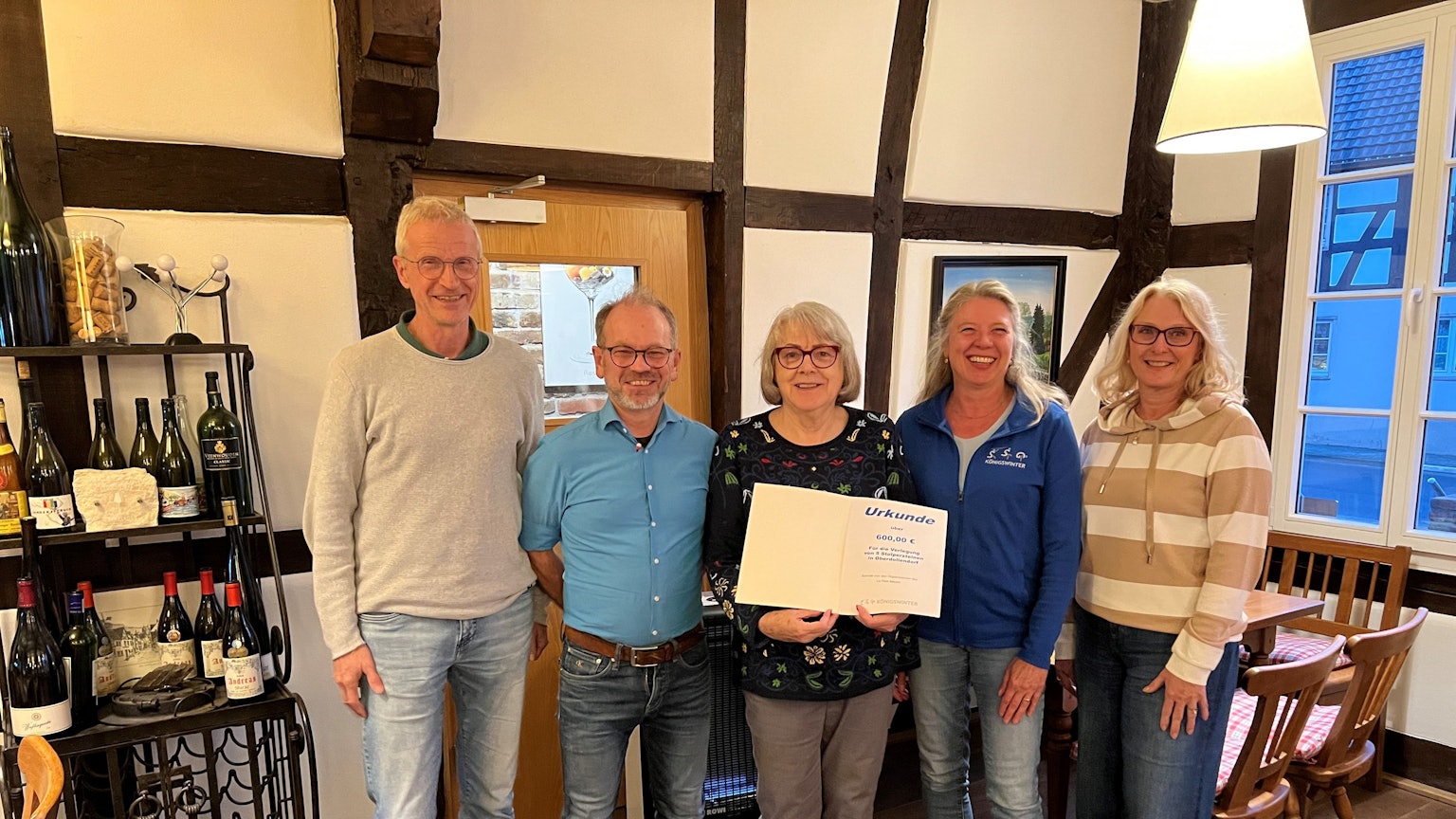 Zwei Männer und drei Frauen mit einer Urkunde, an der Seite steht ein Regal mit Weinflaschen.
