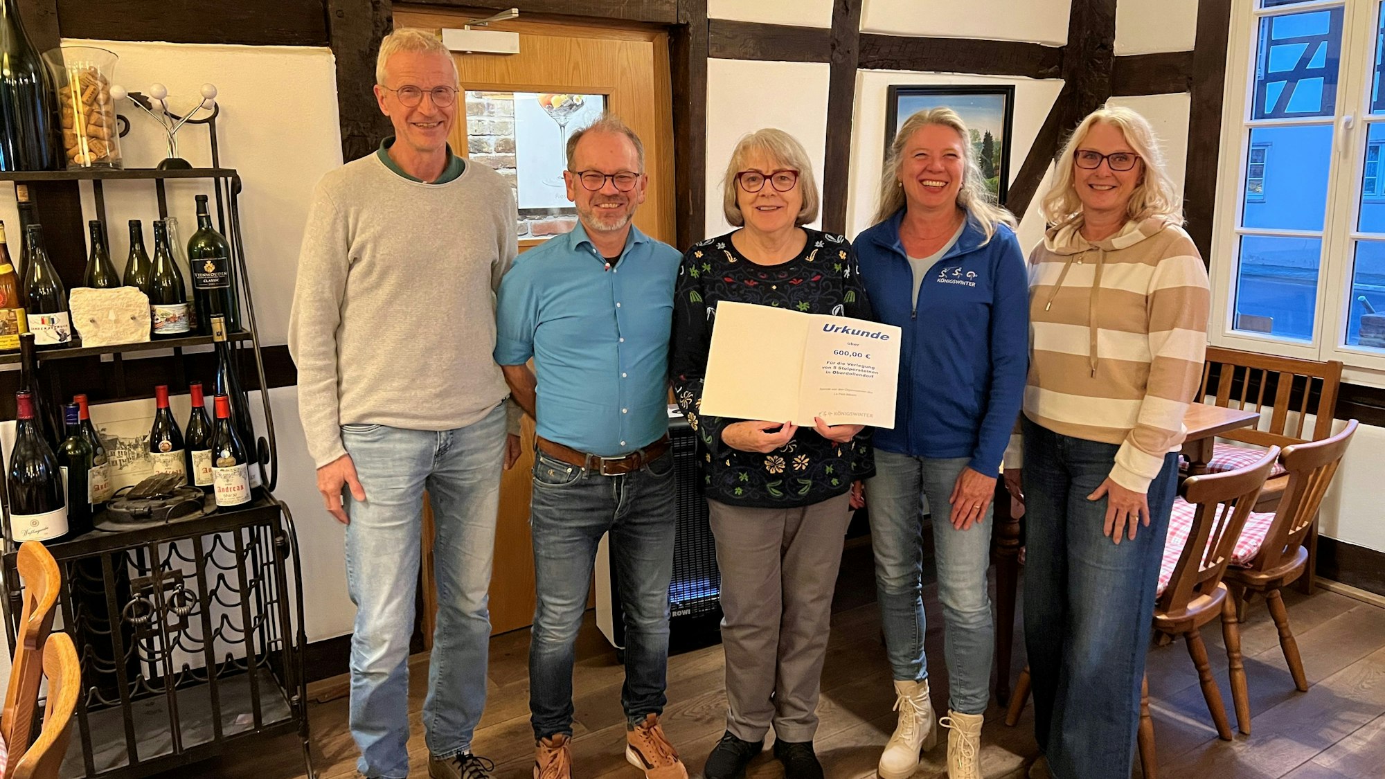 Zwei Männer und drei Frauen mit einer Urkunde, an der Seite steht ein Regal mit Weinflaschen.
