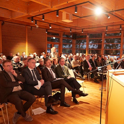 Der Vorsitzende der Jury steht am Rednerpult, vor ihm sitzen die Gäste.