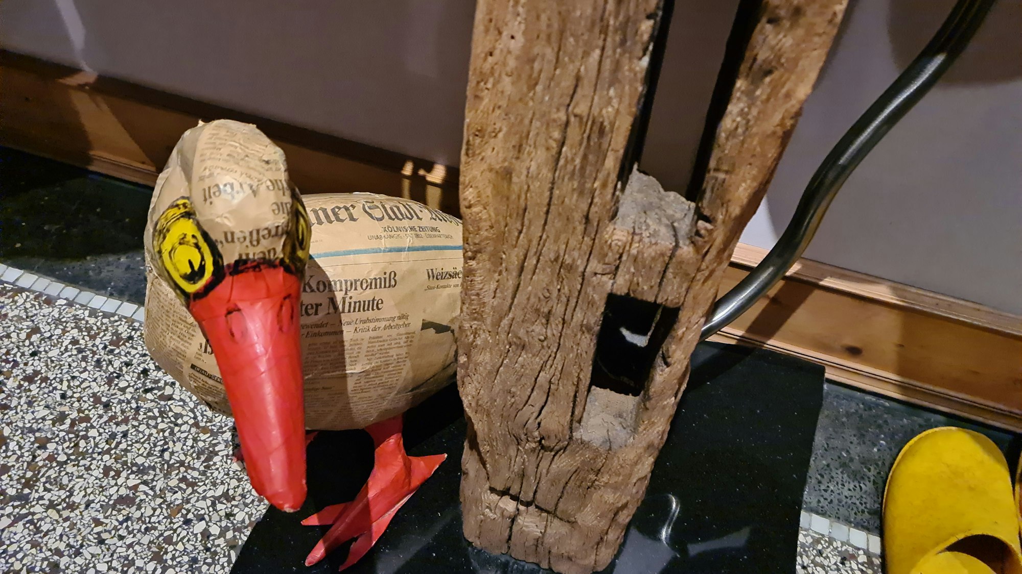 Eine aus Zeitungspapier gefertigte Ente lugt hinter einem Holzbalken hervor.