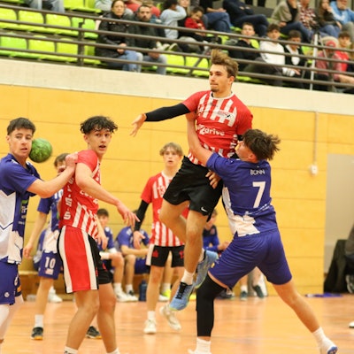 Der geworfene Ball eines Handballers, der in der Luft steht, landet im Gesicht des Gegenspielers. Ein weiterer Gegenspieler hat sich beim Werfer eingehakt.