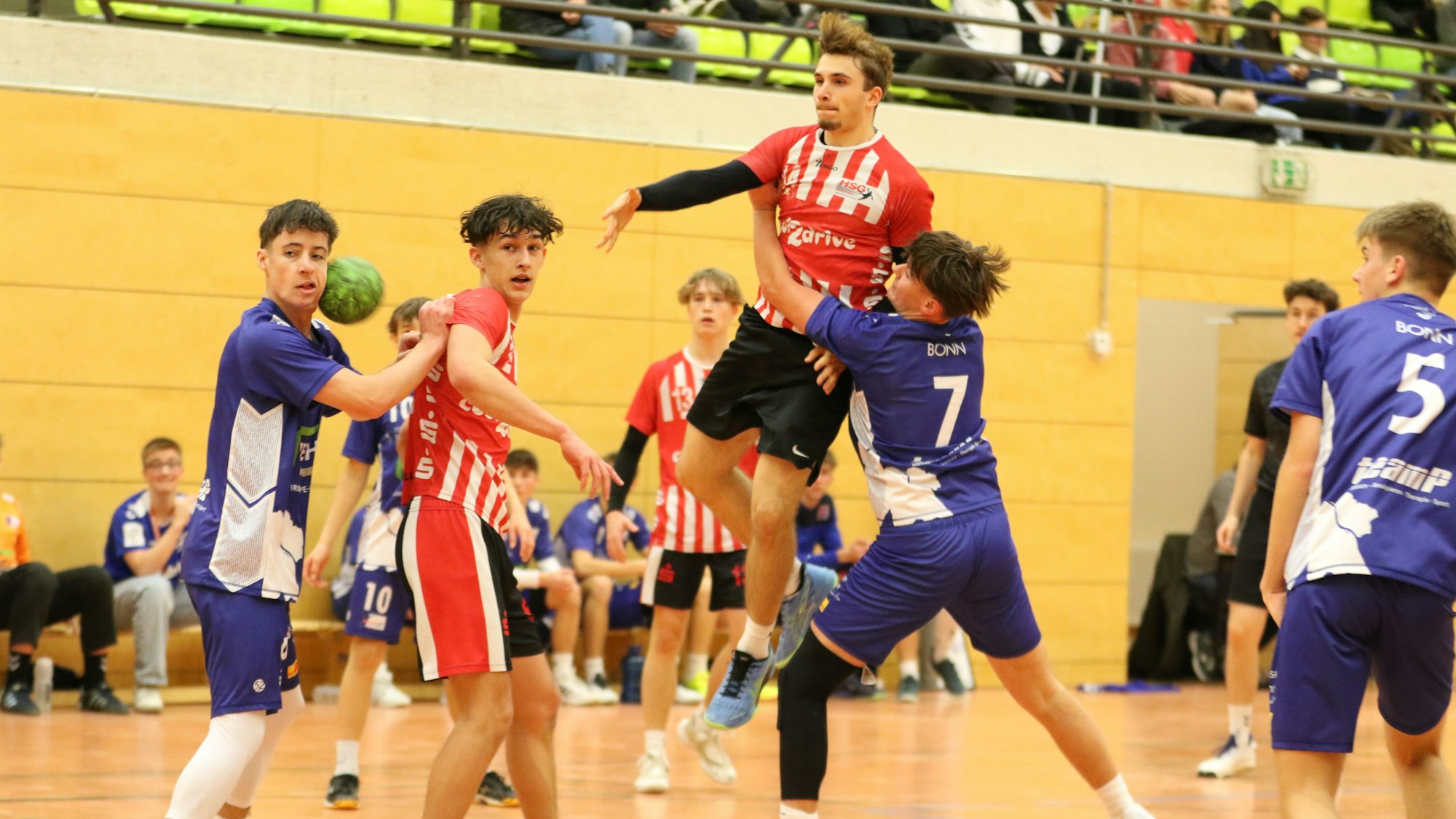Der geworfene Ball eines Handballers, der in der Luft steht, landet im Gesicht des Gegenspielers. Ein weiterer Gegenspieler hat sich beim Werfer eingehakt.