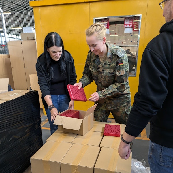 Hauptmann Nadine L. nimmt ein Geschenk, das in rotes Papier verpackt ist, und steckt es in ein Paket. Eine Zivil-Beschäftigte der Bundeswehr hält die Kiste auf.